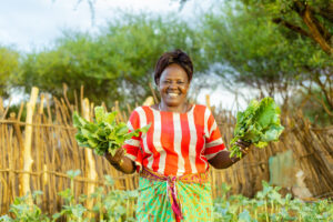 Donna del Kenya con vestito a righe rosso e bianco mostra sorridente le insalate da lei coltivate