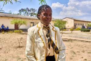 Pearl, una ragazza dello Zimbabwe, indossa la sua uniforme scolastica con giacca e cravatta e sorride di fronte all'edificio scolastico
